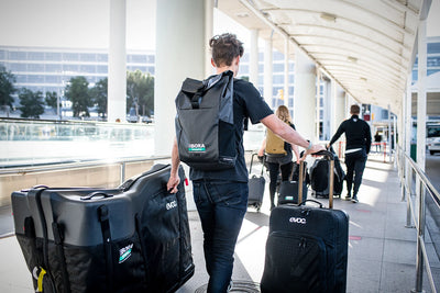 BORA-hansgrohe cycling team rider walking through airport wearing EVOC duffel backpack and rolling Road Bike Bag Pro and World Traveler luggage