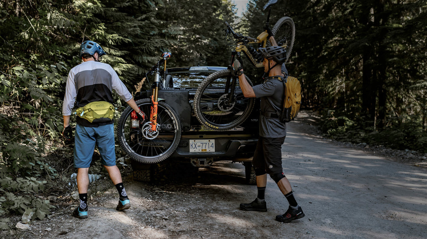 Mountain bikers wearing EVOC hip pack and hydration pack next to truck with tailgate pad