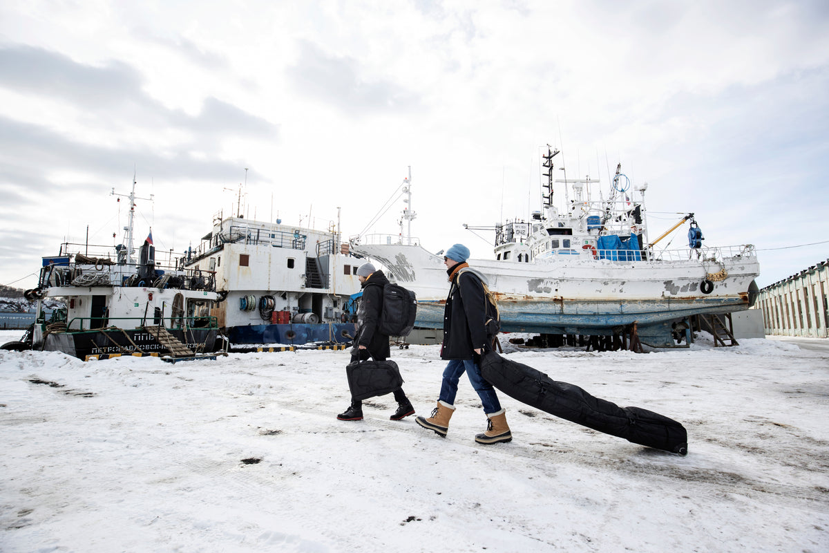 EVOC Snow Gear Roller, Gear Backpack, and Gear Bag being used to transport ski and snowboard equipment in a snowy shipyard